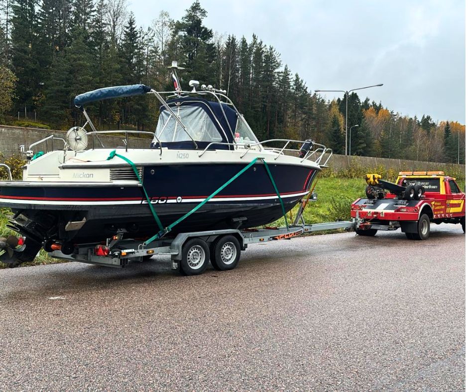 veneen kuljetusta trailerilla ja edessä punainen hinausauto. Taustalla metsää.